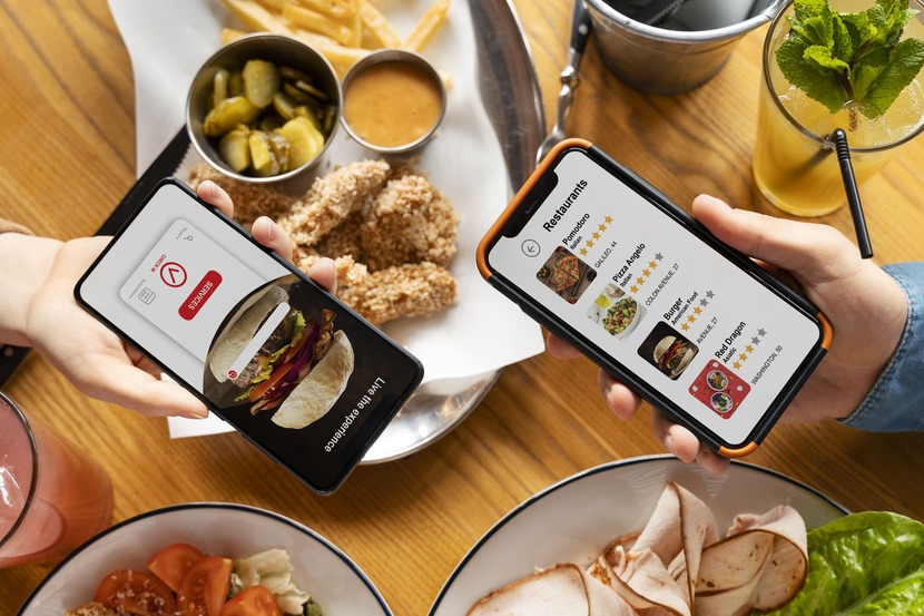 Dois celulares abertos em aplicativos de delivery ao fundo de muita comida
