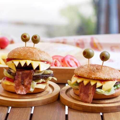Dois hambúrgueres criativos para Halloween: pão com gergelim, queijo cortado em dentes, bacon como "língua" e azeitonas espetadas no topo imitando olhos de monstro. Estão sobre tábuas de madeira, com fundo desfocado e claro.