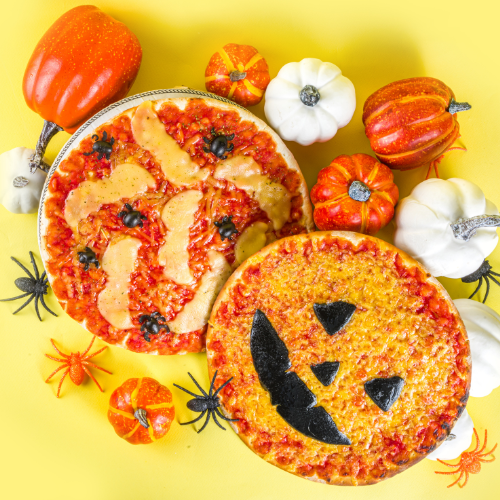 Duas pizzas temáticas de Halloween: uma com queijo em formato de múmia e aranhas de azeitona, outra como abóbora com rosto. Pequenas abóboras decorativas e aranhas plásticas estão espalhadas em um fundo amarelo.