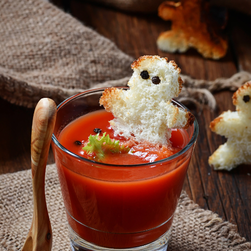 Um copo de vidro com sopa de tomate vibrante, decorado com um "fantasma" de pão torrado e dois olhos de pimenta. Há também um raminho de folhas verdes e uma colher de pau ao lado. O fundo é de madeira rústica e tecido.