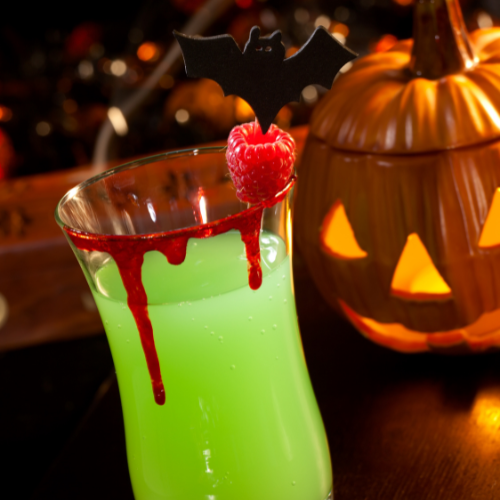 Um coquetel verde vibrante, com calda vermelha escorrendo pela borda, decorado com uma framboesa e um palito de morcego preto. Ao fundo, uma abóbora iluminada e luzes bokeh criam uma atmosfera festiva de Halloween.