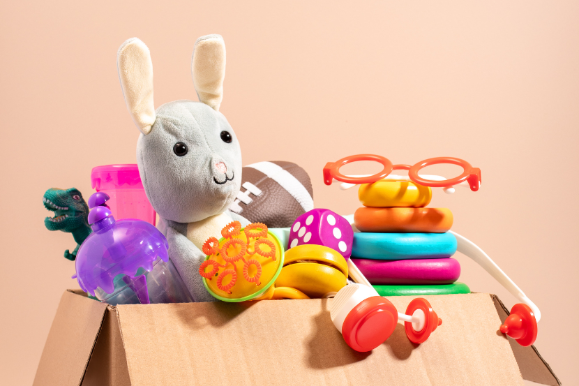 Uma caixa de papelão transborda brinquedos coloridos: um coelho de pelúcia, um dinossauro verde, bolas, um anel empilhável e óculos de brinquedo. O fundo é liso e bege.
