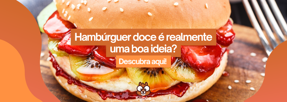 Close de um hambúrguer doce feito com pão de gergelim, recheado com frutas como morango, kiwi e banana, cobertas por calda vermelha. O texto sobreposto diz: "Hambúrguer doce é realmente uma boa ideia? Descubra aqui!". Palavra-chave: hamburguer doce. Saipos.