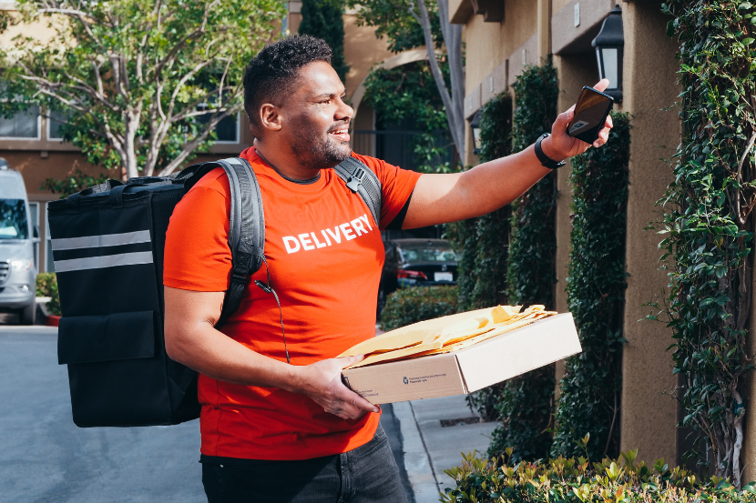 Um entregador barbudo, vestindo camiseta vermelha com "DELIVERY" e mochila preta, segura uma caixa com envelopes enquanto tira uma foto com o celular. Ele está em frente a uma residência com folhagens verdes e um carro branco ao fundo.