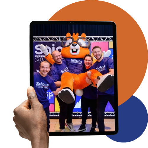 Mão segurando um tablet que exibe uma foto dos fundadores da Saipos com o mascote da empresa, o esquilo Allien.