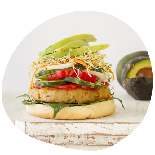 imagem de um hambúrguer vegano com em uma tábua de madeira e no fundo à um abacate e elementos claros para trazer os elementos visuais do produtos e toda a atenção para ele. o hambúrguer é feito com proteína de soja e folhas verdes.