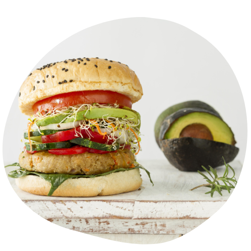 imagem de um hambúrguer vegano com em uma tábua de madeira e no fundo à um abacate e elementos claros para trazer os elementos visuais do produtos e toda a atenção para ele. o hambúrguer é feito com proteína de soja e folhas verdes.