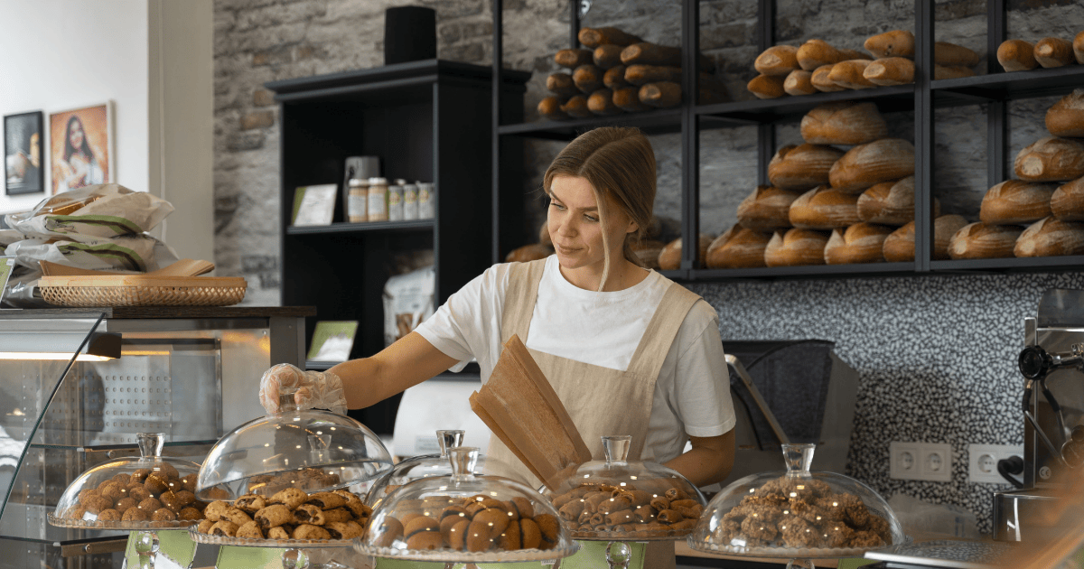 Interior de padaria com balcão repleto de doces sob cúpulas de vidro, pães nas prateleiras e atendente de avental bege. Ambiente acolhedor, bem iluminado e organizado.