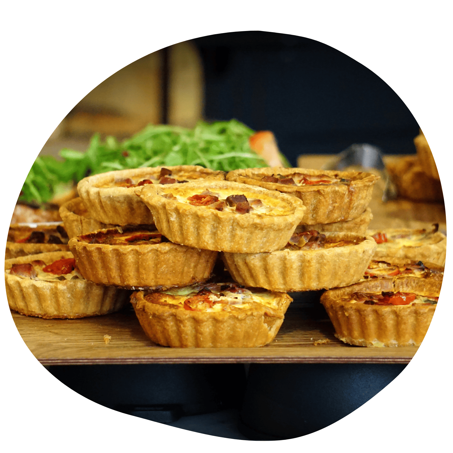 Várias mini quiches ou tortinhas salgadas empilhadas sobre balcão de madeira. Recheio dourado com queijo e bacon, com folhas verdes ao fundo.