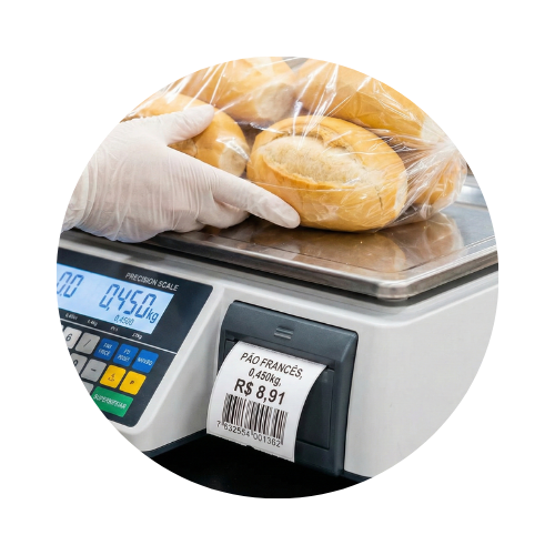 Imagem circular de duas maçãs vermelhas sobre balança digital em um comércio. Uma mão aponta para o visor enquanto um cliente segura uma cesta de compras ao fundo.