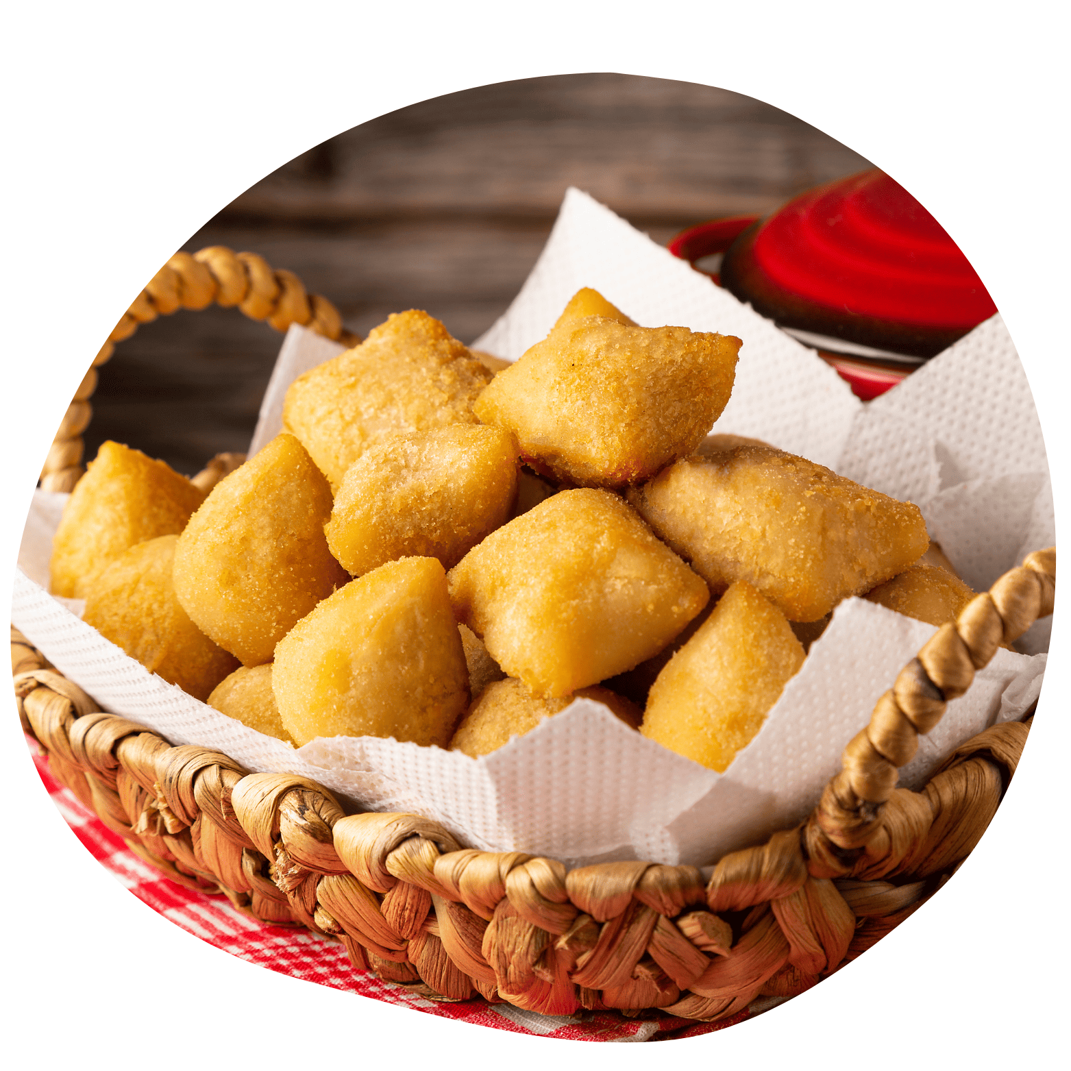 Cesta de vime com papel toalha repleta de rissoles fritos. Fundo rústico com detalhe de panela vermelha.
