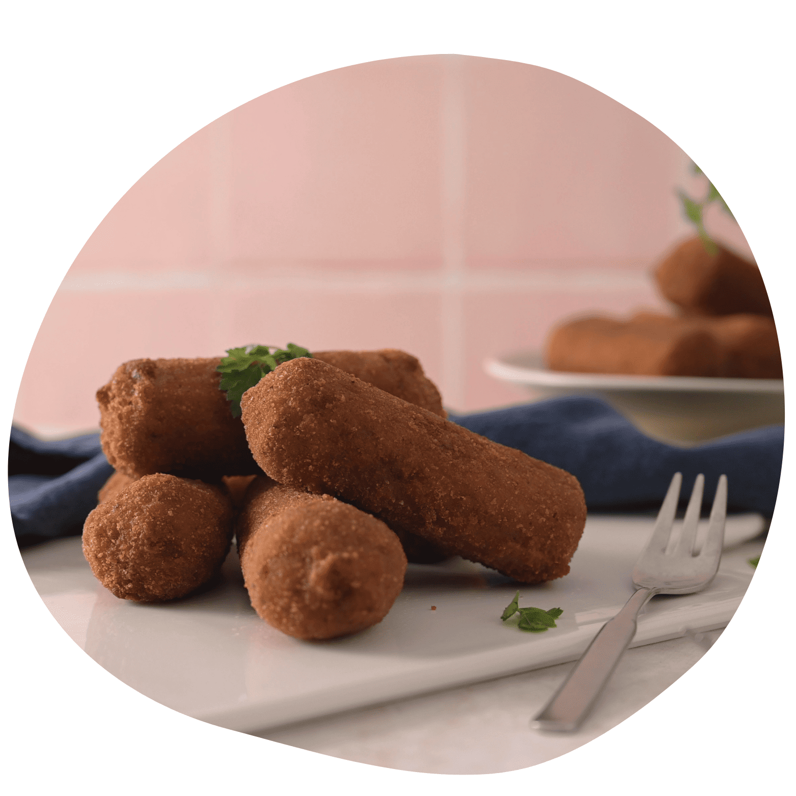 Prato branco com croquetes de carne fritos e um garfo ao lado sobre guardanapo azul. Fundo rosa claro com ramos de salsa decorativos.
