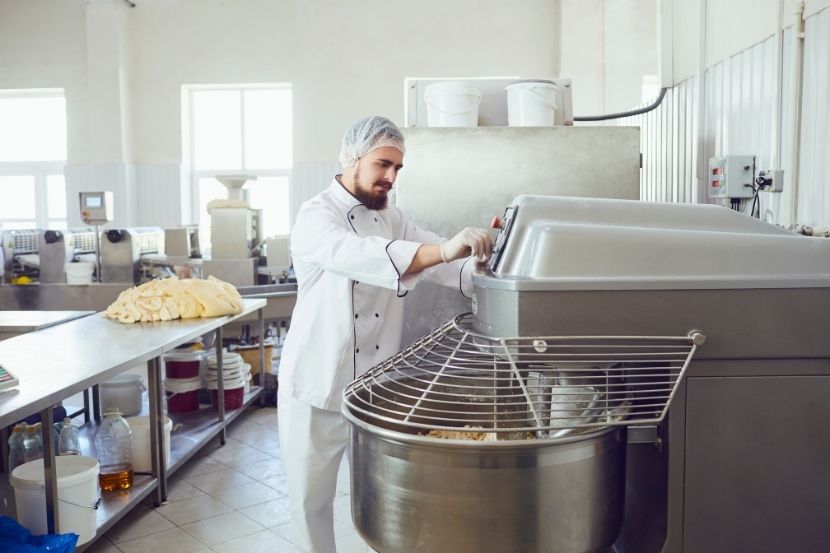 Homem de barba, usando dólmã branca e rede no cabelo, opera uma batedeira industrial de grande porte em uma cozinha profissional. Ao lado, uma bancada de metal com massa de pão.