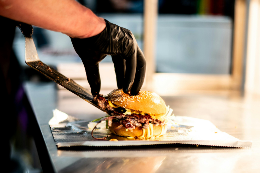 mão com luva preta coloca carne dentro do hamburguer com uma espátula.
