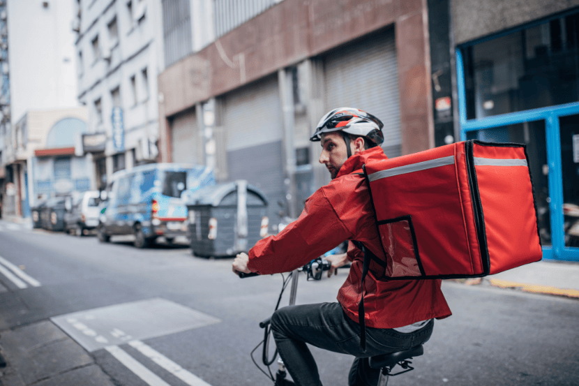 Entregador em bicicleta em rua urbana, visto de perfil. Ele usa jaqueta vermelha, capacete e carrega uma mochila térmica de entrega também vermelha. Ao fundo, prédios e veículos estacionados.