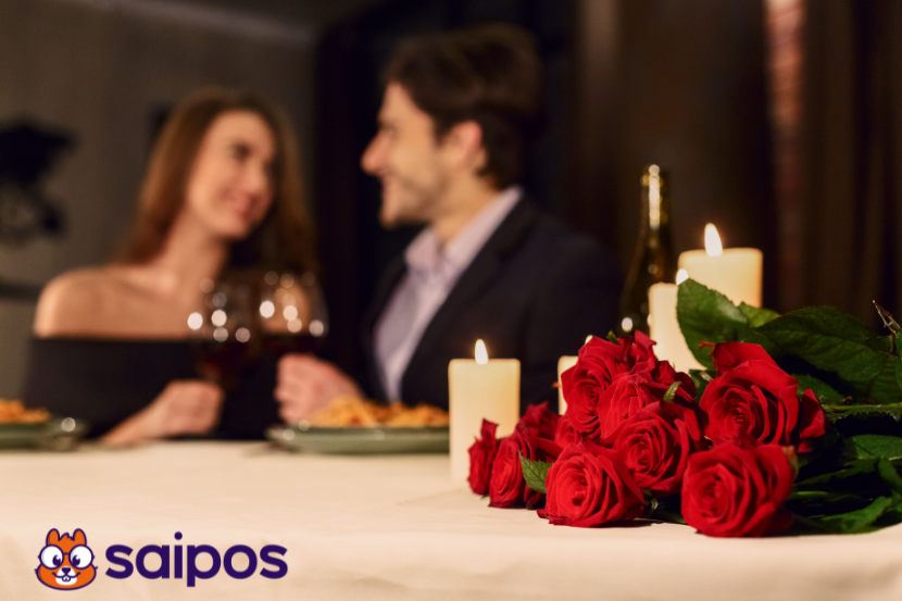 homem e mulher ao fundo em mesa de restaurante bebendo vinho desfocados. À frente, velas brancas e rosas vermelhas sobre toalha branca.