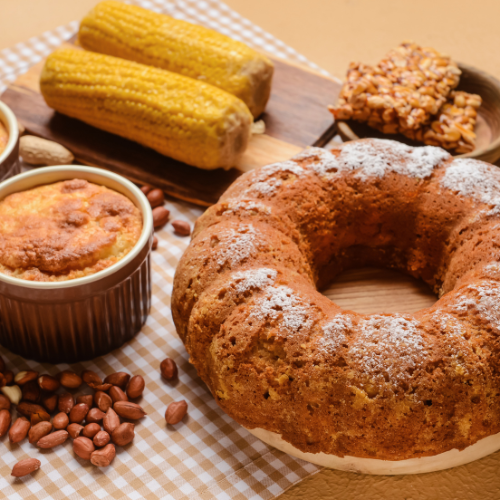 Mesa com toalha quadriculada, bolo de fubá, queijadinha, espigas de milho e pé de moleque. 