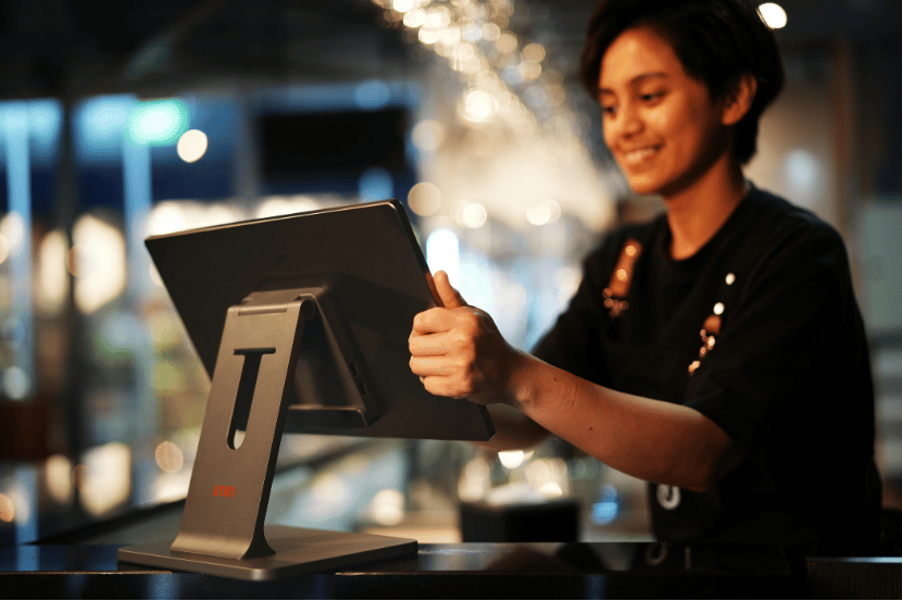 Atendente sorridente de uniforme preto opera terminal de ponto de venda em tablet sobre balcão de restaurante, fazendo sinal positivo com polegar para cima