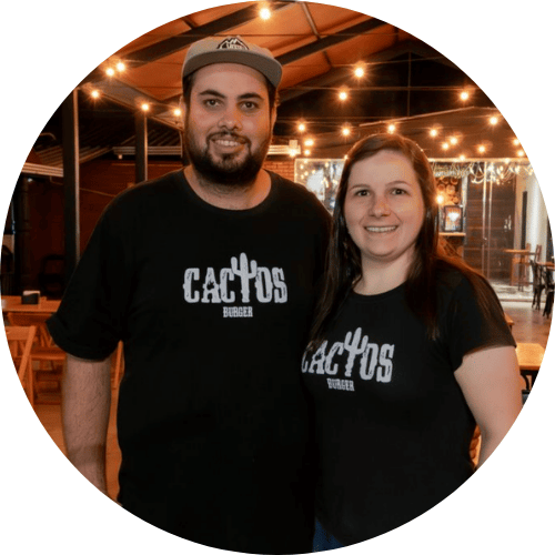 Casal sorridente vestindo camisetas pretas com logotipo Cactos Burger, em ambiente de restaurante decorado com varal de luzes amarelas, homem de boné e barba à esquerda, mulher de cabelos castanhos à direita