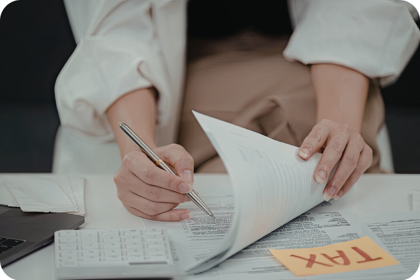 imagem mostra braços com mangas brancas segurando papéis sobre mesa de madeira marrom cheia de outras folhas, além de uma caneta dourada e um post-it escrito "tax".
